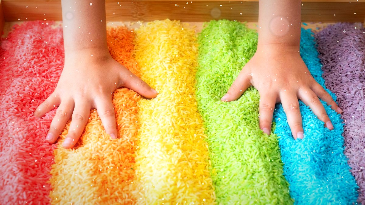 A child's hands running through rows of rainbow-coloured sensory rice